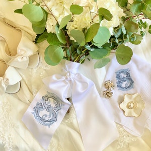 May include: A white bridal bouquet featuring white hydrangeas and eucalyptus foliage, tied with a white satin ribbon adorned with a blue embroidered monogram. The bouquet rests on a white satin fabric with a white lace overlay.