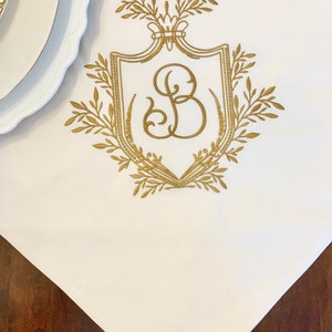 May include: A white linen tablecloth with a gold embroidered crest featuring a stylized letter "B" surrounded by leafy branches.