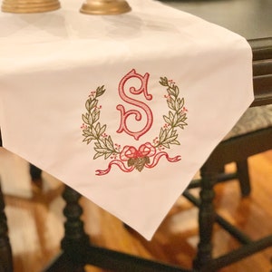 May include: White table runner with an embroidered monogram of the letter "S" in red surrounded by a green wreath with red berries and a red bow with a pine cone.