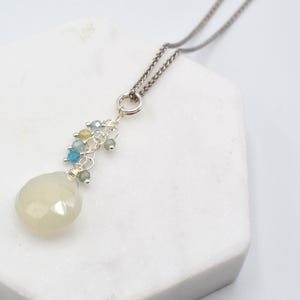 May include: A silver necklace with a pendant featuring a large, faceted, pale yellow gemstone. The pendant is accented with a cluster of small, colourful beads in shades of blue, green, and yellow. The necklace chain is a dark silver colour.