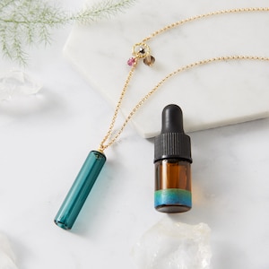 Op de afbeelding: Een gouden ketting met een tealblauwe glazen hanger en kleine edelsteenaccenten. Een klein flesje etherische olie met een tealblauw en groen label is ook afgebeeld.
