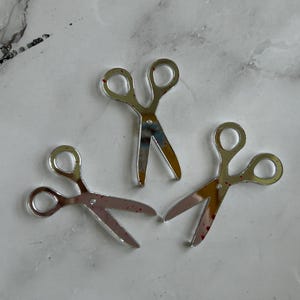 May include: Three silver-colored scissors with open blades are arranged on a light-colored surface. The scissors have a reflective surface and small red spots on the blades. The scissors are a craft or sewing supply.