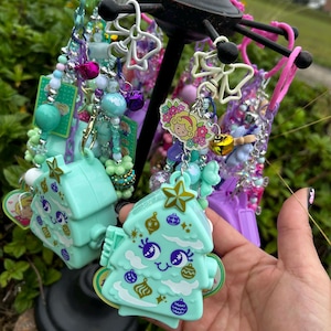 May include: A display of colorful keychains and charms. The keychains feature various designs, including a light blue Christmas tree with a smiling face and decorative ornaments. Other charms include beads, bells, and floral designs.