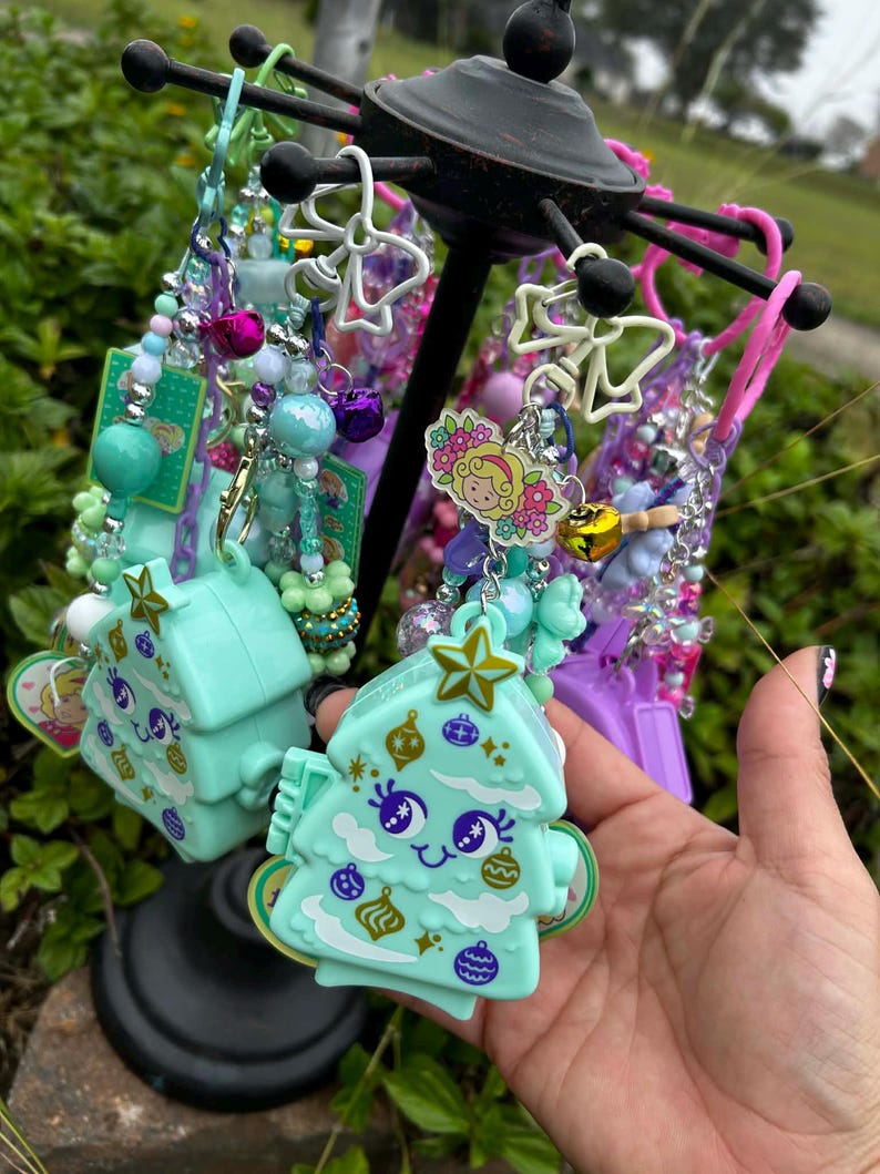 May include: A display of colorful keychains and charms. The keychains feature various designs, including a light blue Christmas tree with a smiling face and decorative ornaments. Other charms include beads, bells, and floral designs.