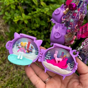 May include: Purple miniature toy with a winter scene inside. One side shows a skier, the other a pink reindeer on a sleigh with presents. The toy is part of a larger charm bracelet with various pink and purple charms.
