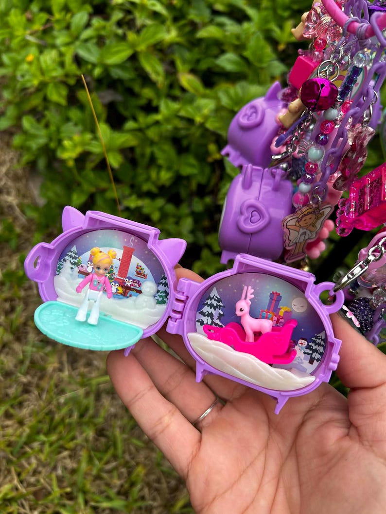 May include: Purple miniature toy with a winter scene inside. One side shows a skier, the other a pink reindeer on a sleigh with presents. The toy is part of a larger charm bracelet with various pink and purple charms.