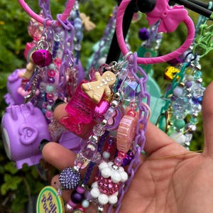 May include: A collection of colorful keychains and charms in shades of pink, purple, and blue. The keychains feature various charms, including a gingerbread man, a bell, and a Polly Pocket logo. The keychains are attached to pink plastic rings.