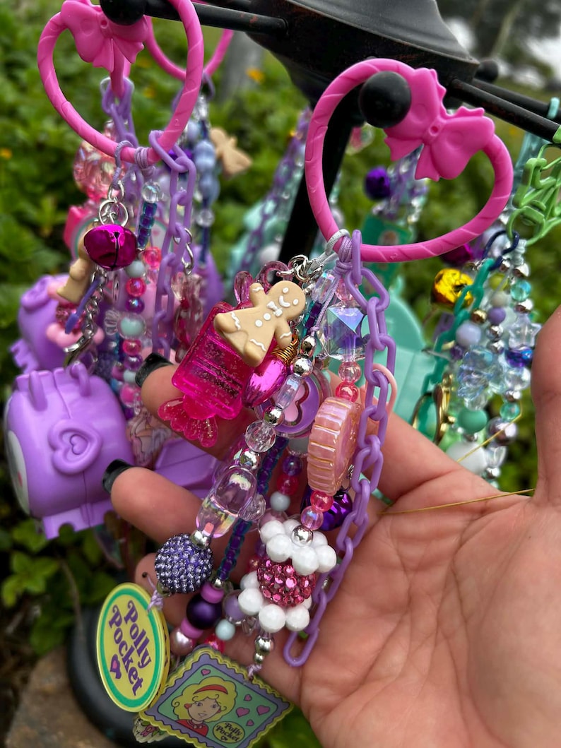 May include: A collection of colorful keychains and charms in shades of pink, purple, and blue. The keychains feature various charms, including a gingerbread man, a bell, and a Polly Pocket logo. The keychains are attached to pink plastic rings.