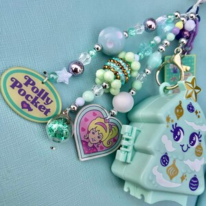May include: A collection of beaded charms and a Polly Pocket Christmas tree-shaped keychain. The charms include a heart-shaped pendant with a cartoon girl, a clear orb with glitter, and a Polly Pocket oval pendant. The color palette is pastel with silver accents.