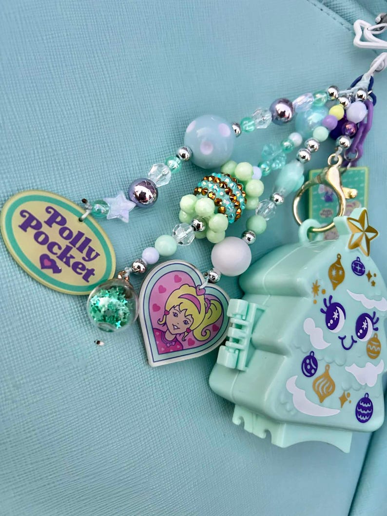 May include: A collection of beaded charms and a Polly Pocket Christmas tree-shaped keychain. The charms include a heart-shaped pendant with a cartoon girl, a clear orb with glitter, and a Polly Pocket oval pendant. The color palette is pastel with silver accents.
