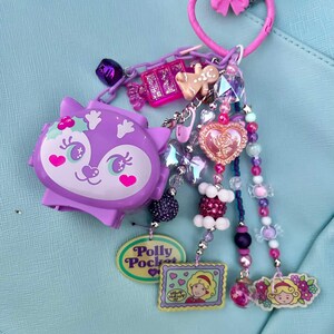 May include: A purple Polly Pocket charm with a variety of decorative beads and charms. The charm features a purple cat-like figure, a gingerbread man, and a pink heart-shaped charm. The charm is attached to a pink heart-shaped keyring.
