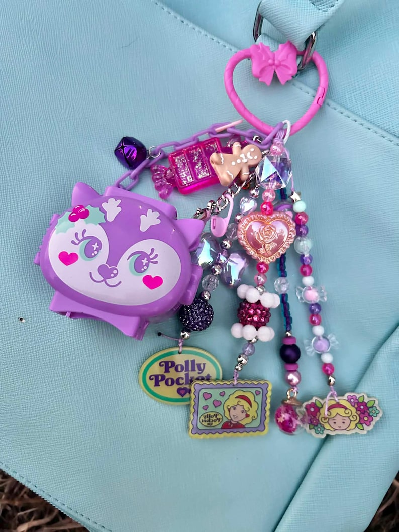 May include: A purple Polly Pocket charm with a variety of decorative beads and charms. The charm features a purple cat-like figure, a gingerbread man, and a pink heart-shaped charm. The charm is attached to a pink heart-shaped keyring.