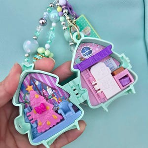 May include: A mint green Christmas-themed miniature house charm, open to reveal a pink Christmas tree and furniture. The charm is attached to a beaded keychain with pastel-colored beads and a decorative bow.