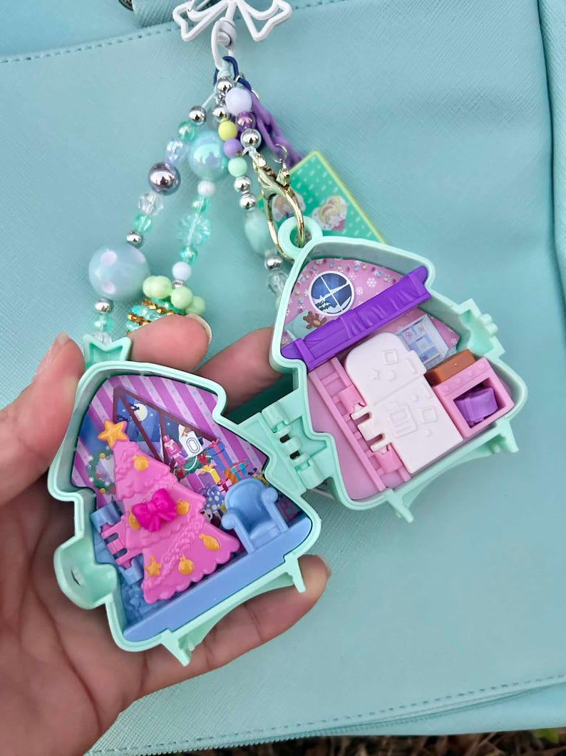May include: A mint green Christmas-themed miniature house charm, open to reveal a pink Christmas tree and furniture. The charm is attached to a beaded keychain with pastel-colored beads and a decorative bow.