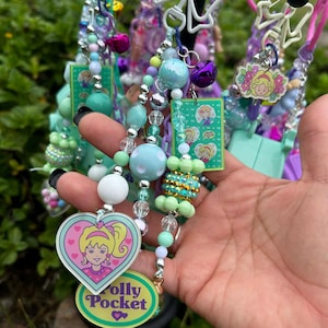 May include: A collection of colorful beaded keychains featuring Polly Pocket designs. The keychains include various beads in shades of blue, green, and silver, with heart-shaped charms and small bell accents. The keychains are attached to a black metal stand.