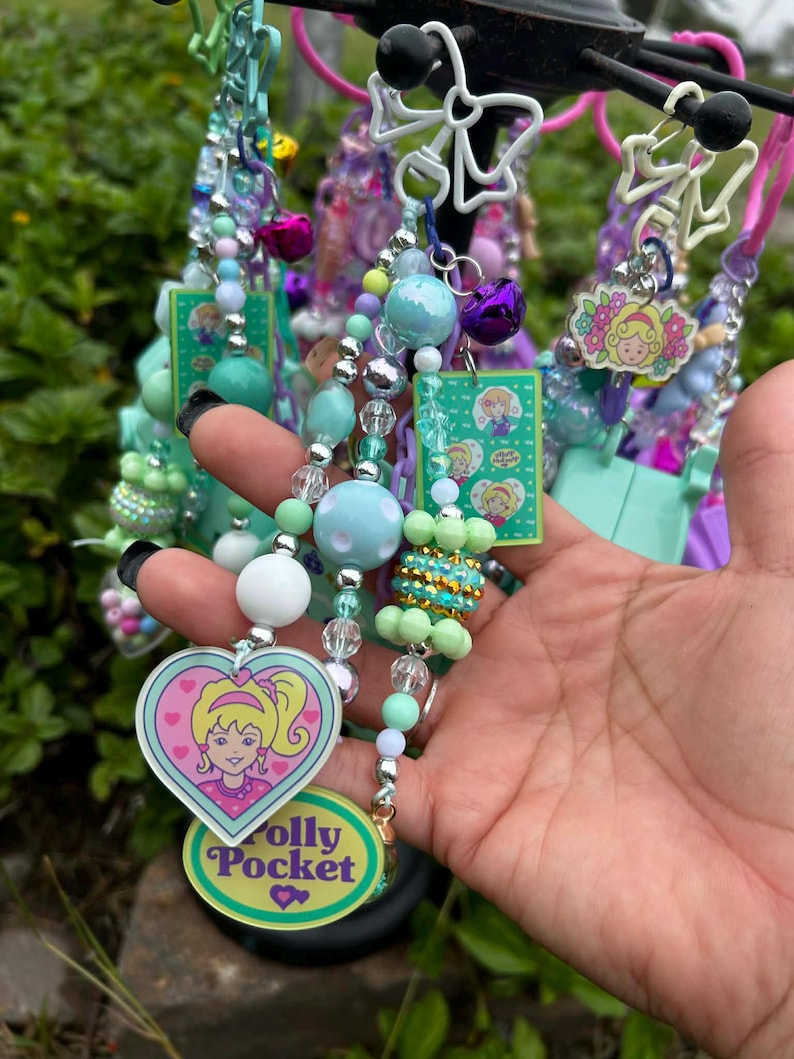 May include: A collection of colorful beaded keychains featuring Polly Pocket designs. The keychains include various beads in shades of blue, green, and silver, with heart-shaped charms and small bell accents. The keychains are attached to a black metal stand.