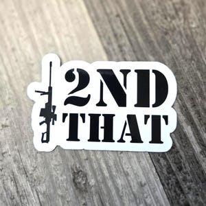 May include: A white sticker with black text and a silhouette of a rifle. The text reads "2ND THAT" in a bold, sans-serif font. The sticker is on a gray wooden surface.