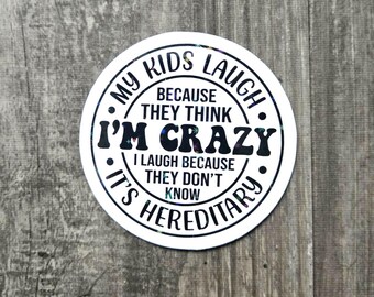 Könnte beinhalten: Ein weißer, runder Aufkleber mit dem Text "MY KIDS LAUGH BECAUSE THEY THINK I'M CRAZY I LAUGH BECAUSE THEY DON'T KNOW IT'S HEREDITARY" in schwarzer, fetter Schrift. Der Aufkleber hat einen schwarzen Rand und einen grauen Holzmaserungshintergrund.