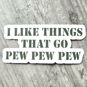 May include: A white sticker with the text "I LIKE THINGS THAT GO PEW PEW PEW" in dark green, stencil-style lettering. The sticker is on a gray, wood-grain surface.