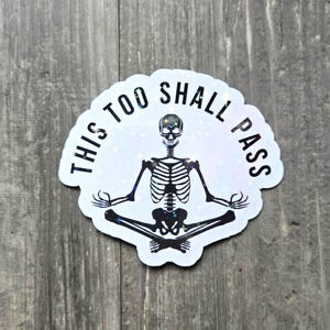 May include: A white sticker with a holographic effect featuring a meditating skeleton. The text "THIS TOO SHALL PASS" is printed around the skeleton. The skeleton is in a seated yoga pose.