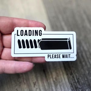 K&ouml;nnte beinhalten: Ein wei&szlig;er Aufkleber mit schwarzem Rand. Er zeigt die W&ouml;rter "LOADING" und "PLEASE WAIT..." und eine Grafik einer Ladeleiste mit schwarzen vertikalen Linien und einem schwarzen Rechteck.