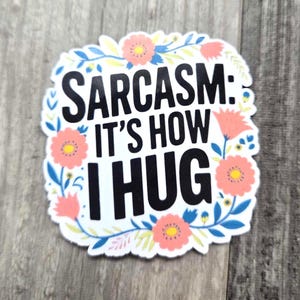 May include: White sticker with a floral border and the text "Sarcasm: It's how I hug" in black.