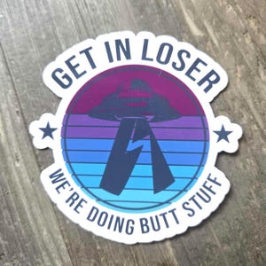 May include: A sticker with a retro design featuring a purple and blue color scheme. The sticker has the text "GET IN LOSER WE'RE DOING BUTT STUFF" around a graphic of a UFO. Two white stars are on either side of the design.