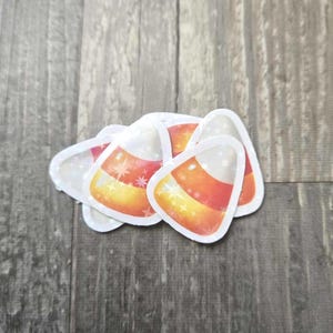 May include: A cluster of candy corn stickers. Each sticker features the classic tri-color design of white, yellow, and orange, with a starburst pattern. The stickers are on a wooden surface.