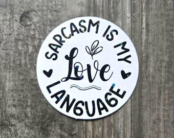 Könnte beinhalten: Ein runder, weißer Aufkleber mit dem Text "Sarcasm is my Love Language" in schwarzer Schrift. Das Wort "Love" ist in einer größeren, kursiven Schriftart geschrieben. Kleine schwarze Herzen und ein Zweig mit Blättern sind ebenfalls enthalten.