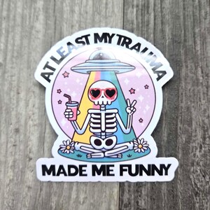 May include: A white sticker with a skeleton sitting cross-legged, holding a pink drink. The skeleton has pink sunglasses and is surrounded by stars and a rainbow. The text reads "At Least My Trauma Made Me Funny".