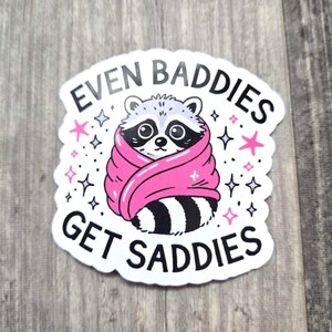 May include: White sticker with a black and white raccoon wrapped in a pink blanket. The sticker says "Even Baddies Get Saddies" with stars around the raccoon.