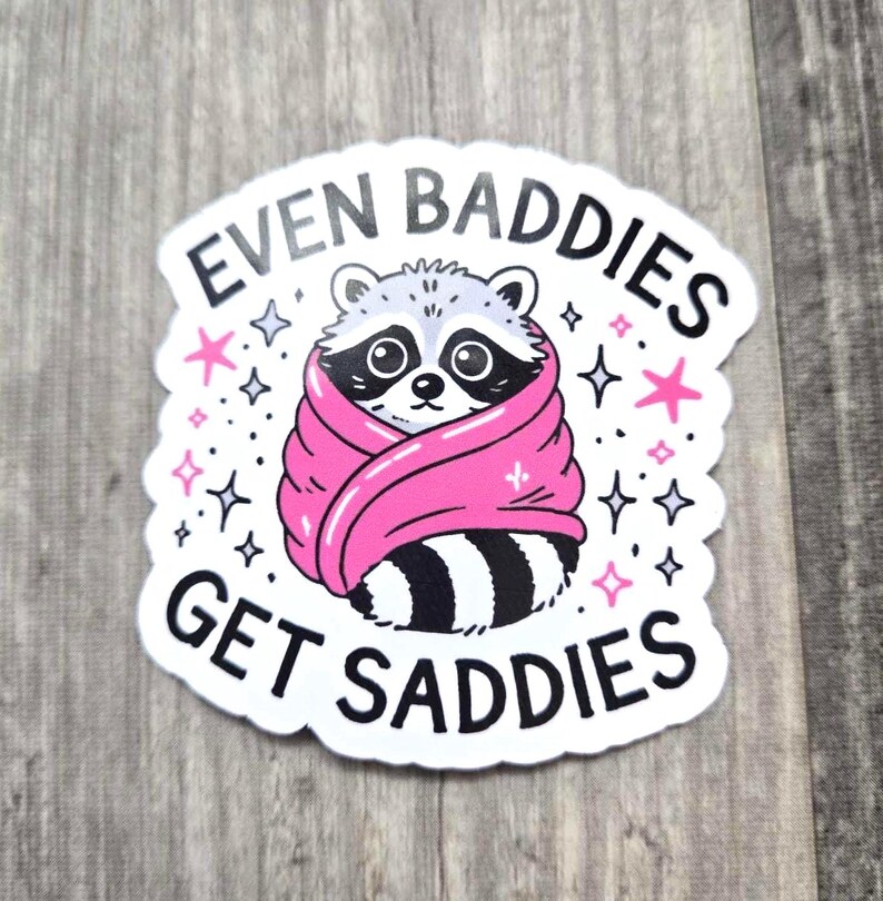 May include: White sticker with a black and white raccoon wrapped in a pink blanket. The sticker says "Even Baddies Get Saddies" with stars around the raccoon.