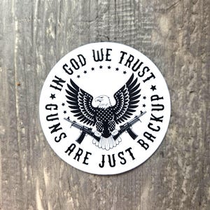 May include: A white circular sticker with a black and white design. The design features an eagle with outstretched wings, two rifles, and the text "IN GOD WE TRUST" and "GUNS ARE JUST BACKUP".