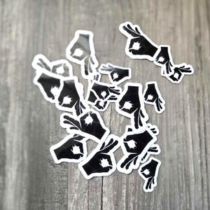 May include: A collection of black and white stickers featuring the "OK" hand gesture. The stickers are scattered on a wooden surface.