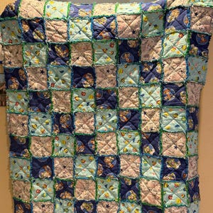 May include: A handmade patchwork quilt featuring a repeating pattern of squares in shades of blue, light blue, and white. The quilt has a rag edge with green stitching, and the squares feature cartoon character designs.