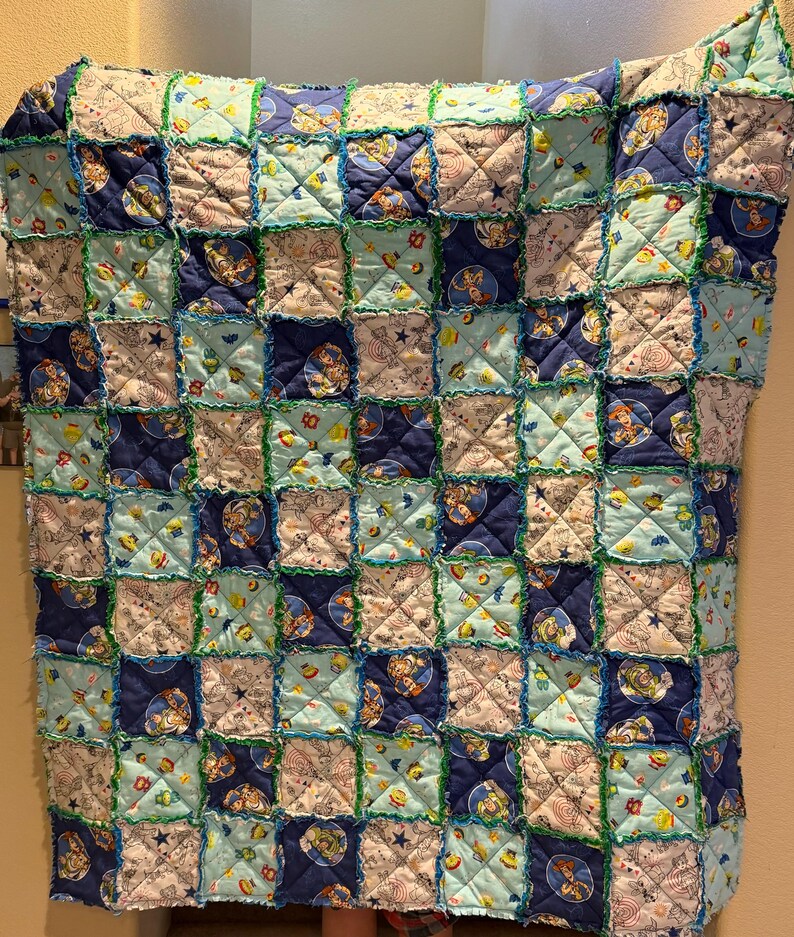 May include: A handmade patchwork quilt featuring a repeating pattern of squares in shades of blue, light blue, and white. The quilt has a rag edge with green stitching, and the squares feature cartoon character designs.