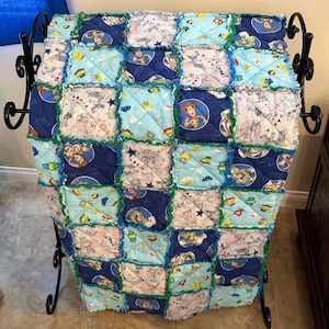 May include: A handmade patchwork quilt featuring a Toy Story theme. The quilt is composed of various squares in shades of blue, light blue, and white, with images of Woody, Buzz Lightyear, and other characters. The edges are trimmed with green fabric.