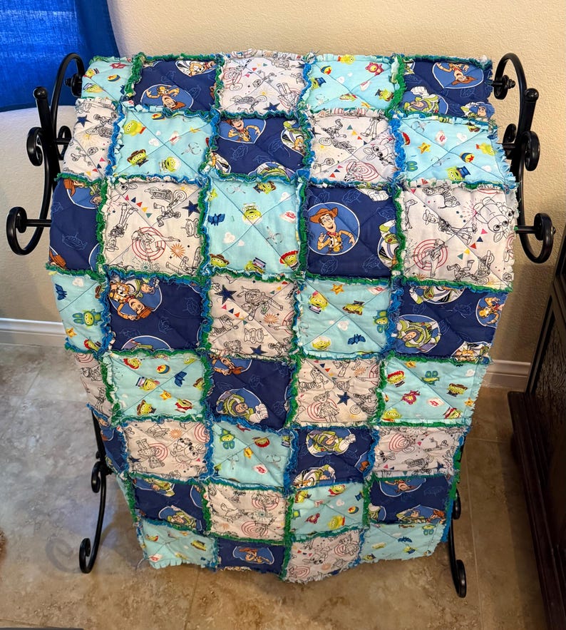 May include: A handmade patchwork quilt featuring a Toy Story theme. The quilt is composed of various squares in shades of blue, light blue, and white, with images of Woody, Buzz Lightyear, and other characters. The edges are trimmed with green fabric.
