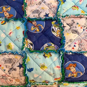 May include: A patchwork quilt featuring squares with Toy Story characters like Woody and the aliens. The squares are in shades of blue, light blue, and white, with green frayed edges. The quilt has a playful, handmade aesthetic.