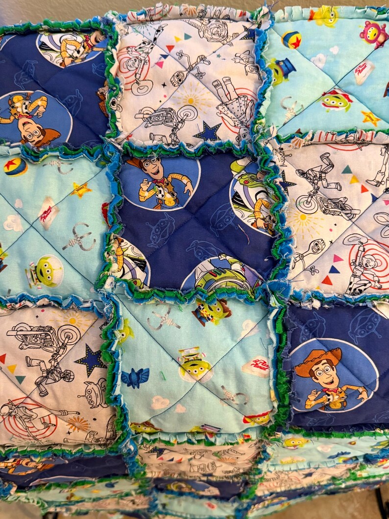 May include: A patchwork quilt featuring squares with Toy Story characters like Woody and the aliens. The squares are in shades of blue, light blue, and white, with green frayed edges. The quilt has a playful, handmade aesthetic.