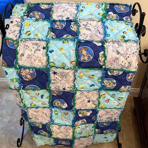 May include: A patchwork quilt featuring squares of various Toy Story themed fabrics in shades of blue, green, and white. The quilt has a ruffled edge and is displayed on a black metal stand.