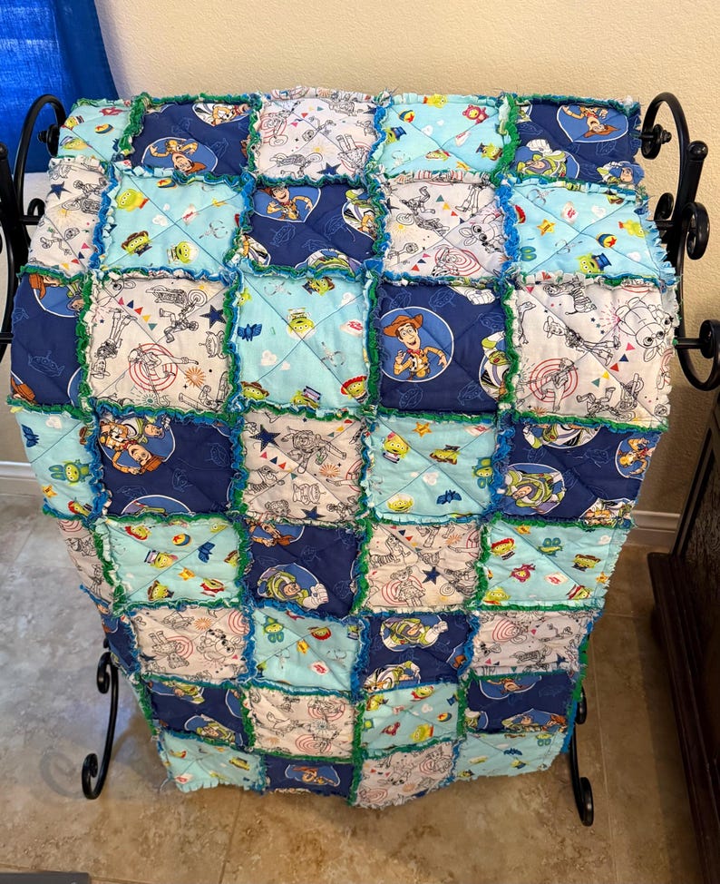 May include: A patchwork quilt featuring squares of various Toy Story themed fabrics in shades of blue, green, and white. The quilt has a ruffled edge and is displayed on a black metal stand.