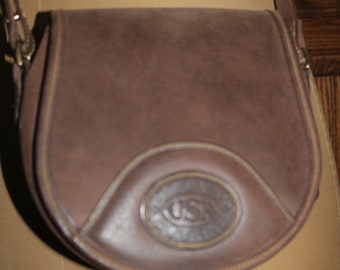 May include: A brown leather satchel bag with a flap closure and a circular logo with the letters "USC" in the center.