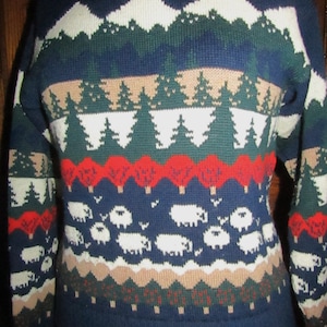 Puede incluir: Un suéter azul, verde, blanco, rojo y marrón con cuello redondo. El suéter presenta un diseño de montañas, árboles y ovejas.