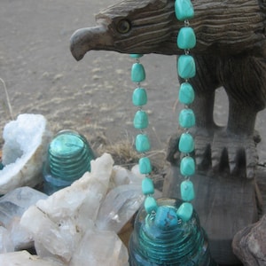 Könnte beinhalten: Eine türkisfarbene Halskette aus Edelsteinen mit einem silbernen Verschluss, präsentiert auf einer Holzadlerskulptur und einer Sammlung von klaren Quarzkristallen.