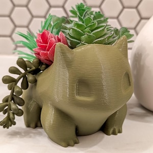 May include: A green 3D printed planter shaped like a Pokemon character, with a small succulent plant growing inside.