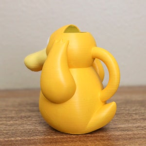May include: A yellow plastic watering can shaped like a cartoon character with a large head and a spout. The watering can has a handle on the side.