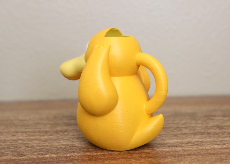 May include: A yellow plastic watering can shaped like a cartoon character with a large head and a spout. The watering can has a handle on the side.