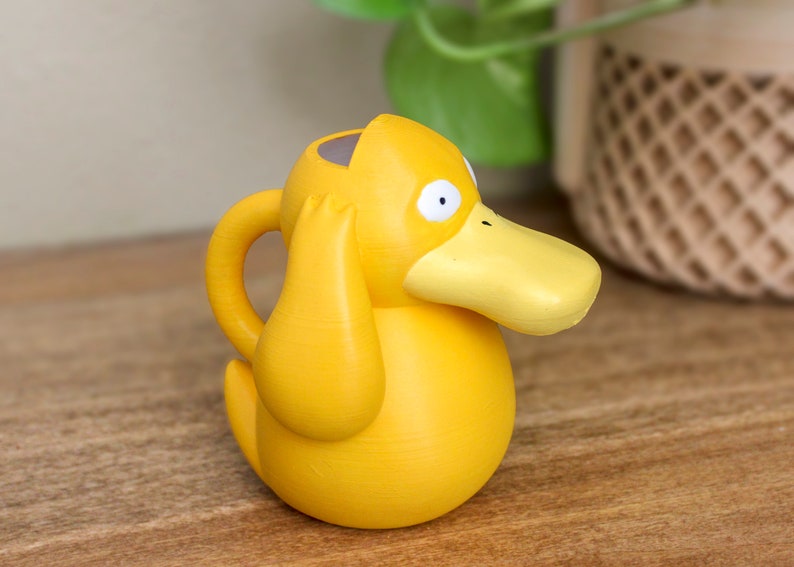 May include: A yellow duck-shaped watering can with a spout and a handle. The duck has large, round eyes and a small beak.