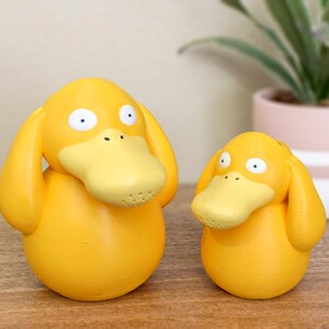 May include: Two yellow duck figurines with closed eyes and outstretched wings. The larger duck is in the foreground and the smaller duck is in the background.